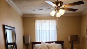 Ein Schlafzimmer mit Bett, Deckenventilator, Nachttisch, Lampe, Stuhl, Fenster mit Vorhängen und Teppich.