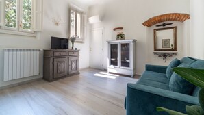 A room with a wooden floor, a blue sofa, a cabinet, a radiator, and a window with a view of trees.