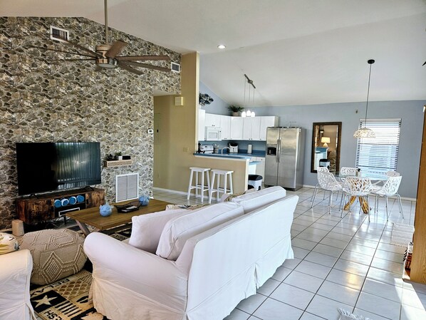 A modern living room with a stone wall, a flat-screen TV, a white sofa, and a kitchen area with white cabinets and a stainless steel refrigerator.