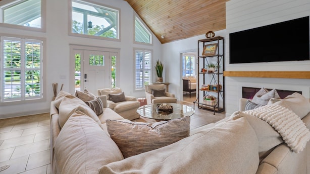 A spacious living room with a high ceiling, large windows, a fireplace, and a flat-screen TV mounted on the wall. - Oleander Cottage! Pet Friendly, Heated Pool, Private Escape, 2 Minutes to Beaches and Restaurants (Saint Simons Island)