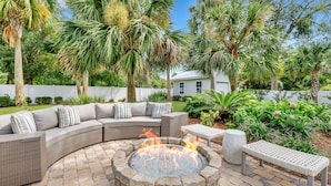 A curved outdoor seating area with a stone fire pit, surrounded by lush greenery and palm trees. - Oleander Cottage! Pet Friendly, Heated Pool, Private Escape, 2 Minutes to Beaches and Restaurants (Saint Simons Island)