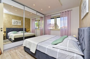 A bedroom with a large mirror, two beds, a window with curtains, and a painting on the wall.