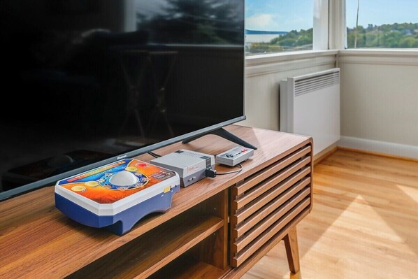 Quarto com uma televisão de tela grande, uma escrivaninha de madeira com gavetas e uma janela com vista para o verde.