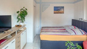 Et soveværelse med seng, skrivebord med tv, en plante og et billede på væggen.