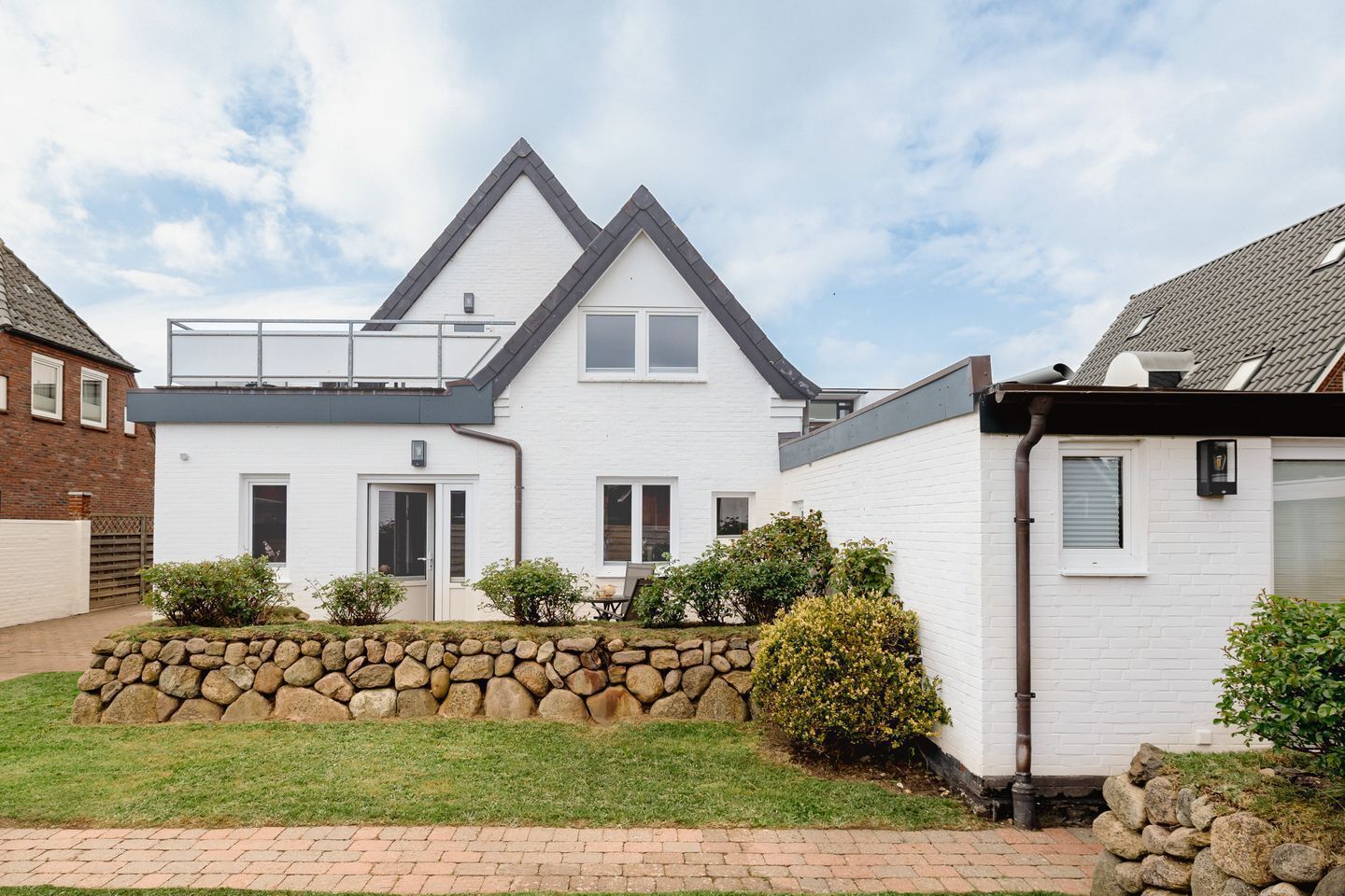 Une maison à deux étages avec un jardin bordé d’un mur en pierre, une allée pavée et un ciel dégagé.
