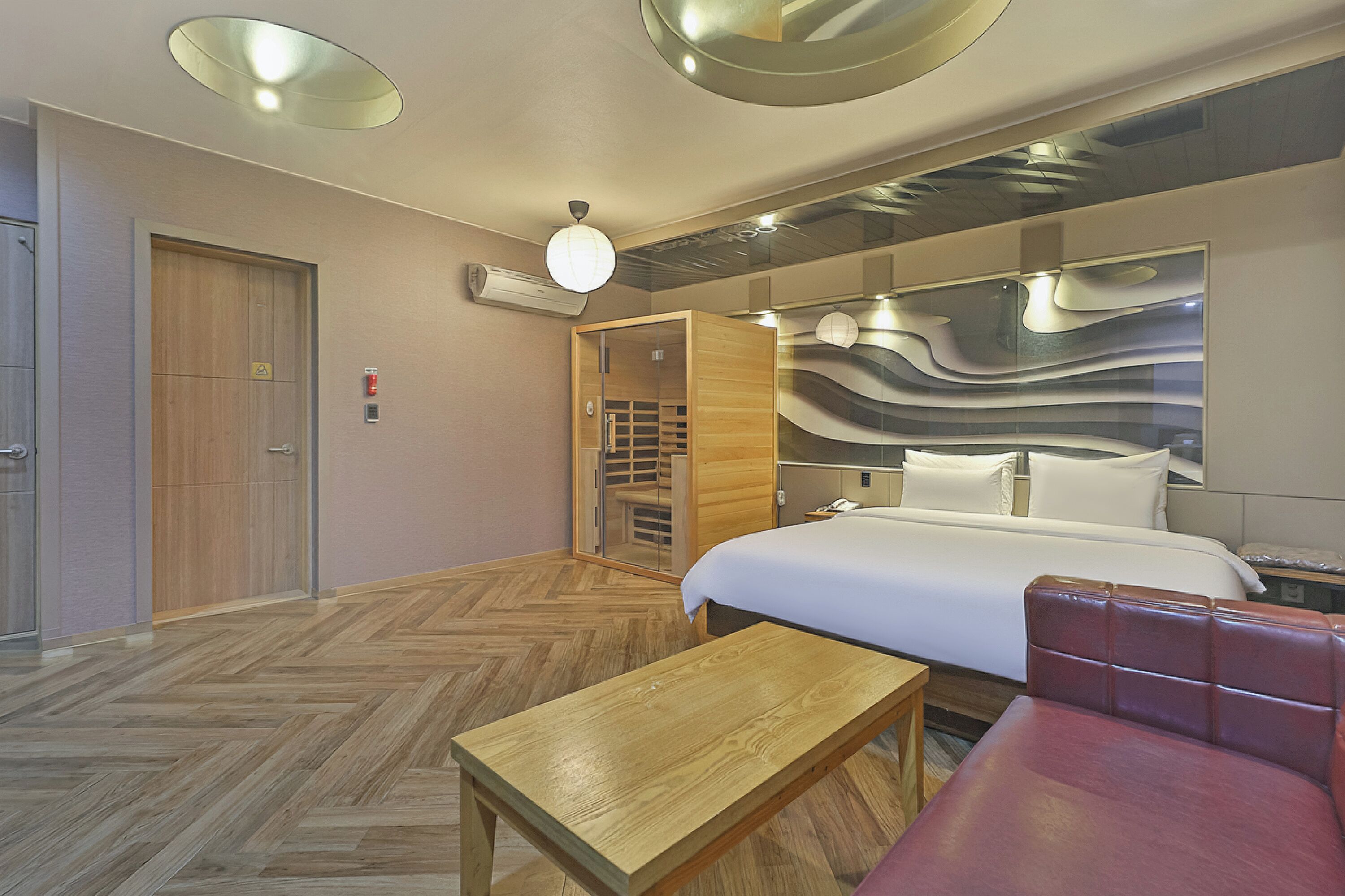 A hotel room with a bed, a wooden bench, a red sofa, a mirror with a wavy design, and a wooden door.