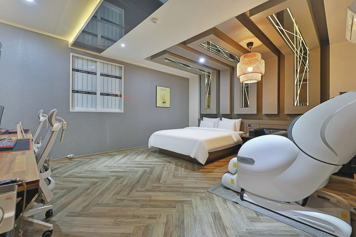 A modern hotel room with a bed, a massage chair, a desk with a computer, and a large window.