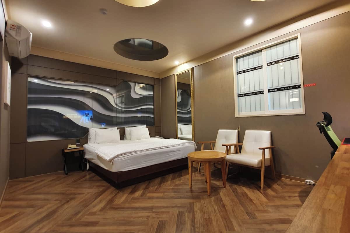 A modern hotel room with a large bed, a small round table, two chairs, a large mirror, and a window with a view of the city.