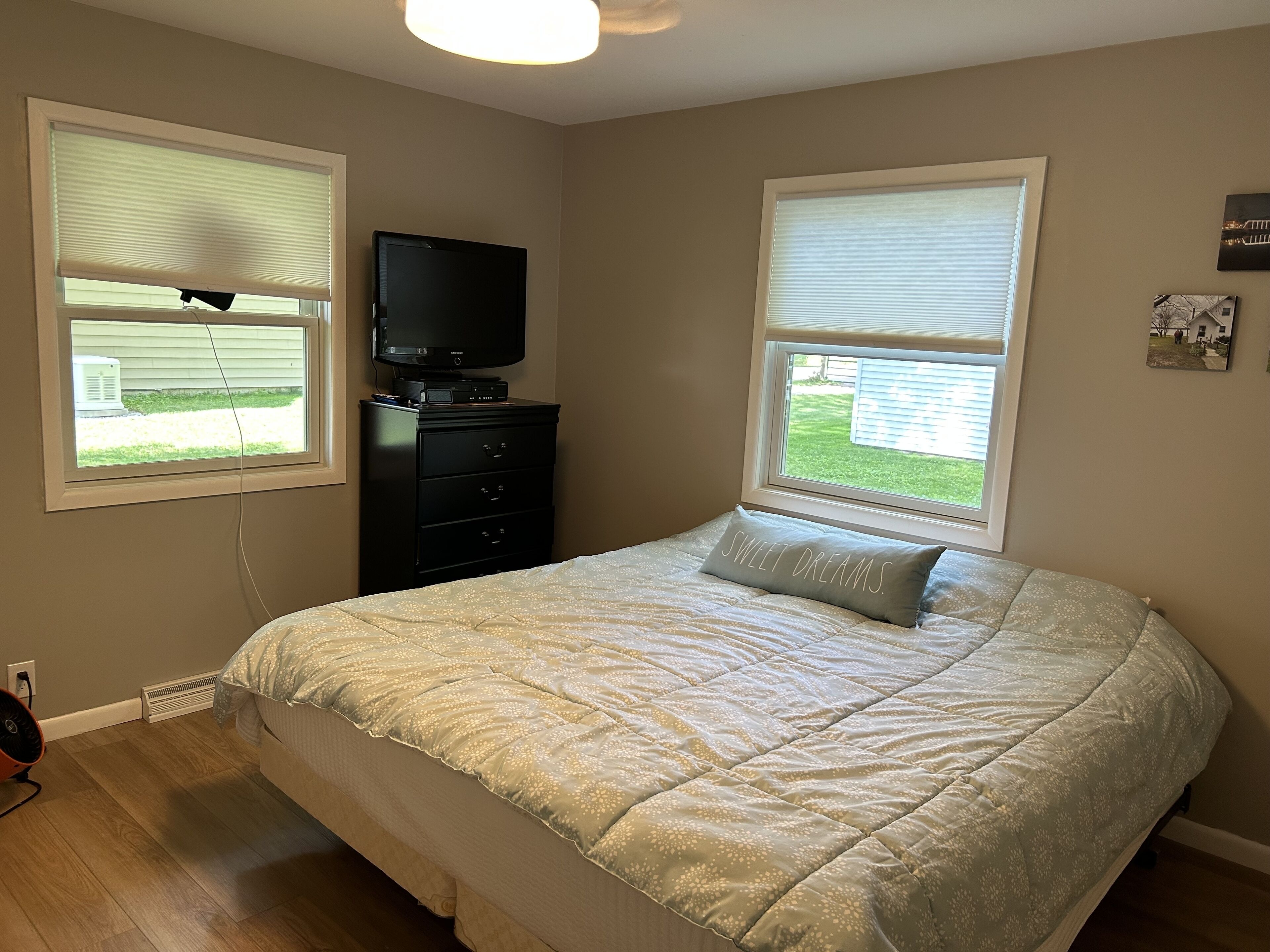 Un dormitorio con cama, cómoda, televisor y dos ventanas con persianas.