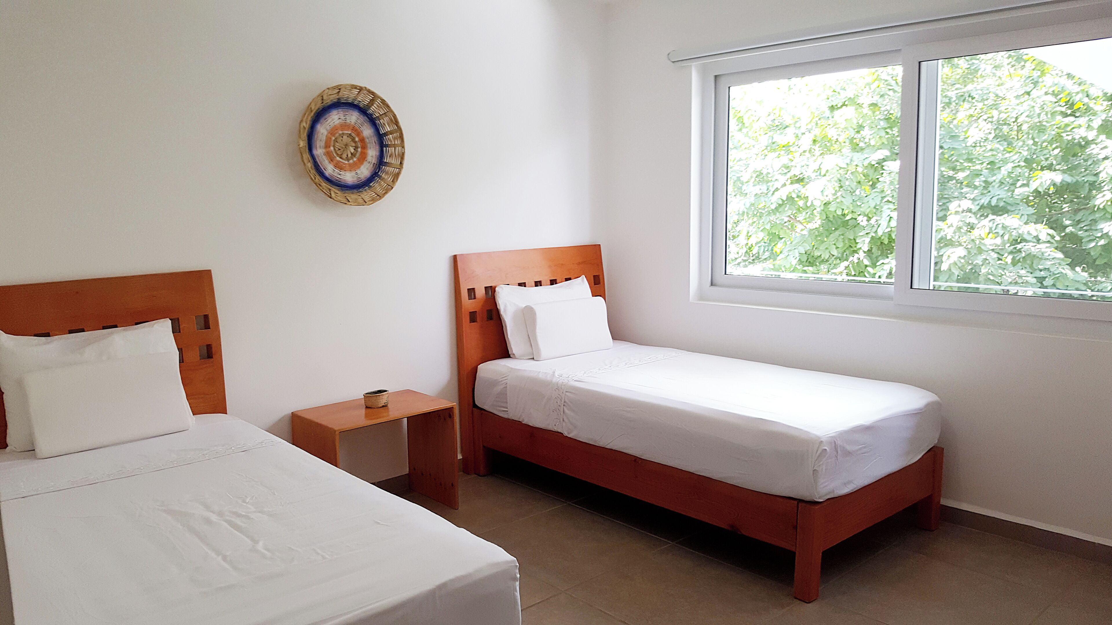 Quarto com duas camas de solteiro, uma pequena mesa de cabeceira com abajur, um adorno na parede e uma janela com vista para o verde.