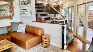 Ein modernes Wohnzimmer mit einem braunen Ledersofa, einem Holztisch und einem einzigartigen Holzschemel. Eine Treppe mit Metallgeländer führt zu einer zweiten Etage.