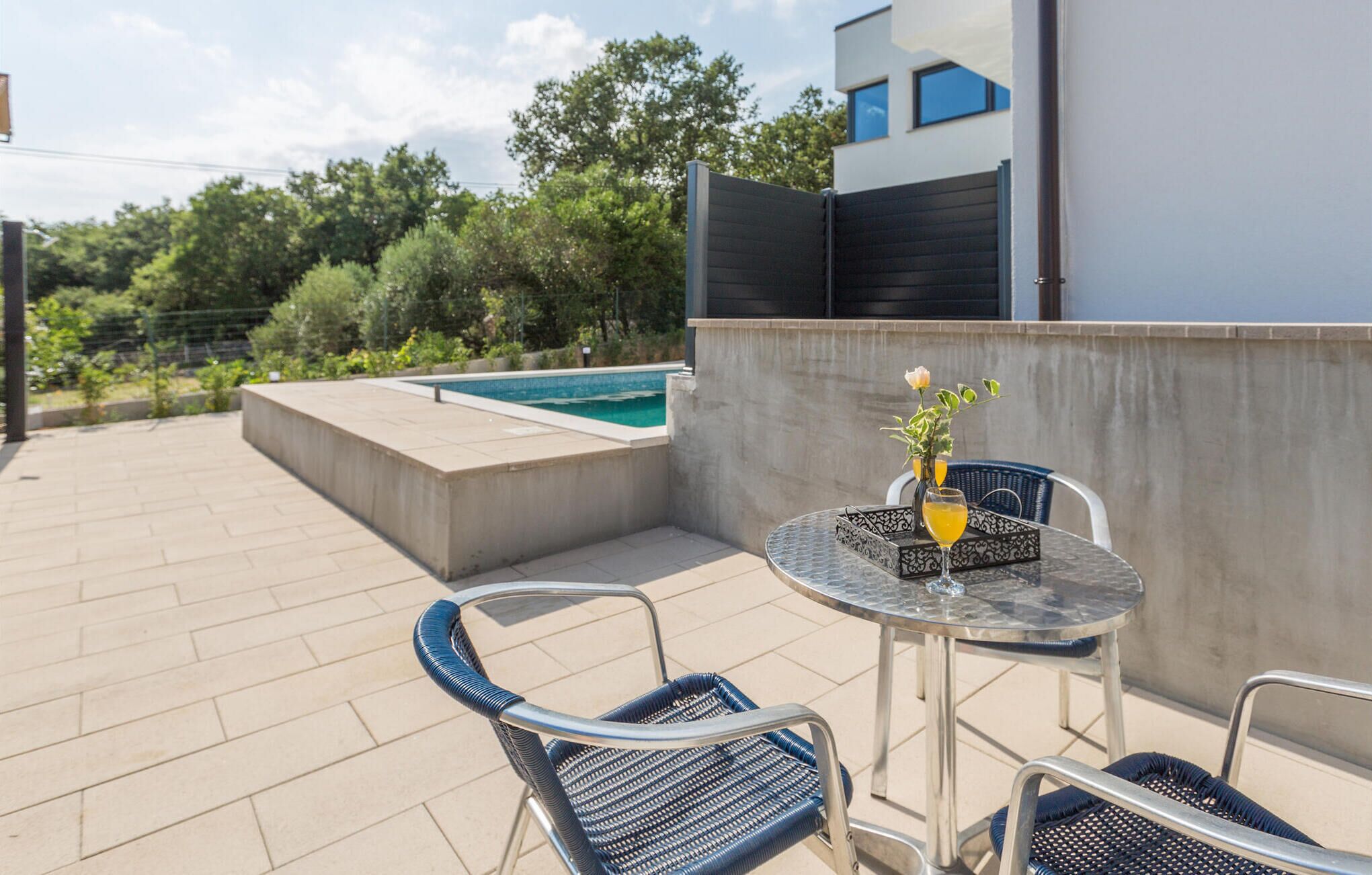 A rooftop terrace with a pool, two chairs, and a table with a vase and drink.