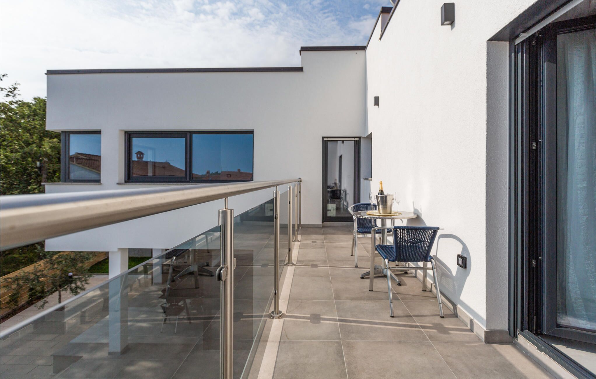 Un balcon extérieur moderne doté d’une rambarde en verre, d’une table avec des chaises et d’une porte donnant sur un espace intérieur.