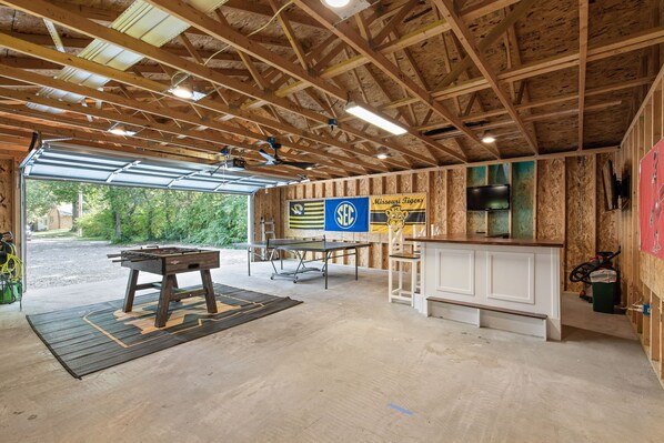 An indoor recreational area with a foosball table, ping pong table, and a pool table. There are wooden walls and a ceiling with exposed beams. A banner for the Missouri Tigers is visible.