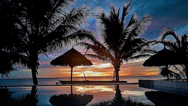 Une vue paisible sur le coucher de soleil, avec des palmiers et un parasol en chaume près d’un bassin aux eaux calmes et réfléchissantes.