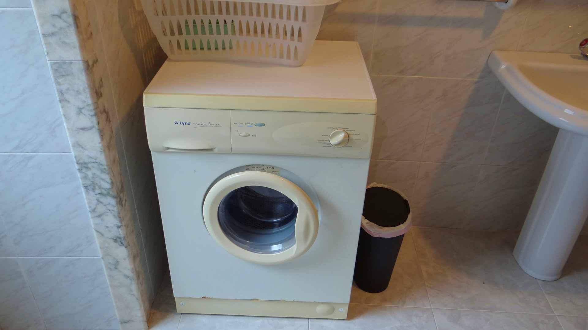 Una lavadora blanca y compacta con puerta frontal, ubicada en un rincón de un baño con paredes y suelo de baldosas.