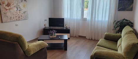 A living room with a flat-screen TV on a stand, a small table with a plant, and two green sofas.