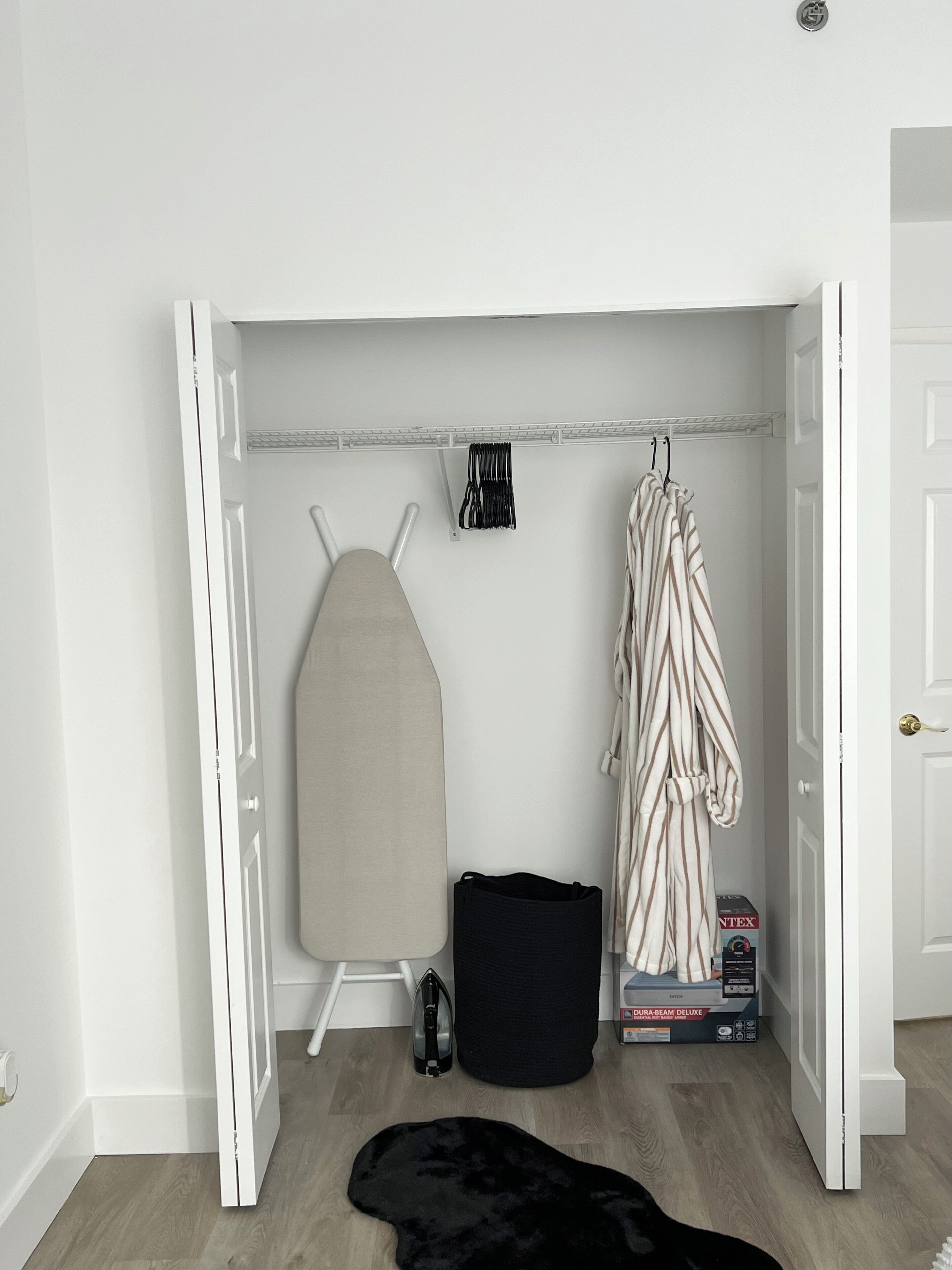 Una habitación compacta con un armario blanco, una tabla de planchar, una percha para ropa, un bolso negro y una alfombra de piel sintética negra.