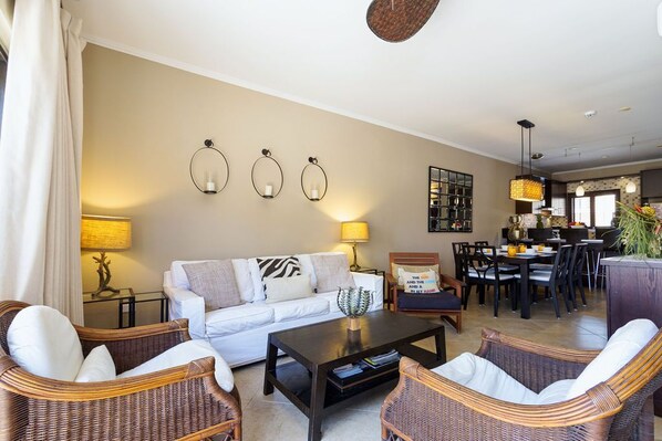 Ein Wohnzimmer mit einem weißen Sofa, Korbstühlen, einem Couchtisch und einem Essbereich mit Tisch und Stühlen.