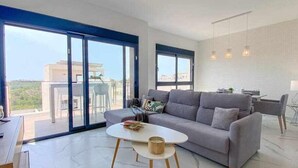 Una sala de estar moderna con un sofá gris, una mesa redonda y un comedor con mesa y sillas. Amplias ventanas ofrecen vistas panorámicas de la ciudad.