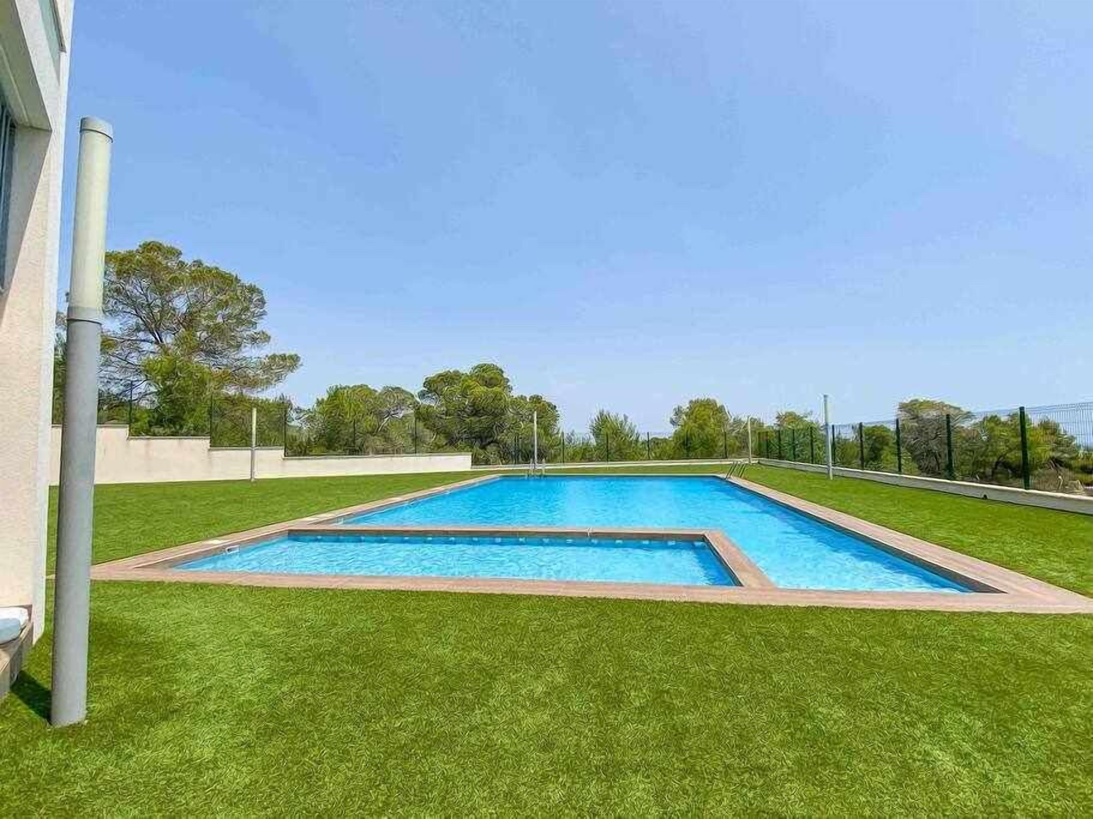 A rectangular swimming pool with a surrounding grassy area, a metal fence, and trees in the background.