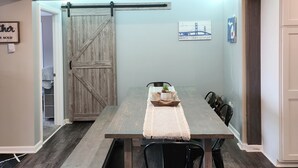 Un comedor con una mesa de madera, sillas negras y un banco. Hay una puerta corrediza de estilo rústico, un cuadro en la pared y una planta pequeña sobre la mesa.
