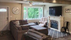 Una sala de estar con sofá, reposapiés y sillones, chimenea, televisor y un reloj en la pared.