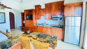A kitchen with wooden cabinets, a granite countertop, a microwave, a refrigerator, and a dining area with chairs. - Beach Front Condo steps from beach (Roatan)