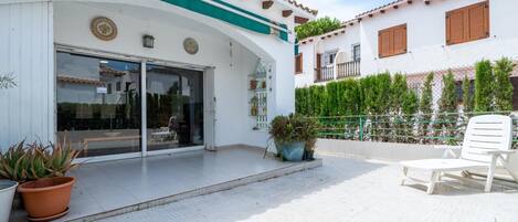 Un edificio con fachada blanca, detalles verdes y un patio empedrado con plantas en macetas y una silla blanca.