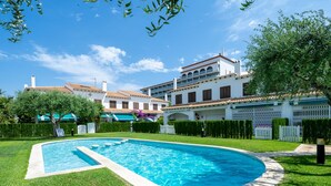 Une piscine entourée d’une pelouse bien entretenue et d’un bâtiment blanc doté d’un toit recouvert de tuiles.