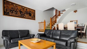 Un salon avec deux canapés en cuir noir, une table basse en bois, un grand tableau au mur, un escalier avec des rampes en bois et un coin repas avec une table et des chaises.
