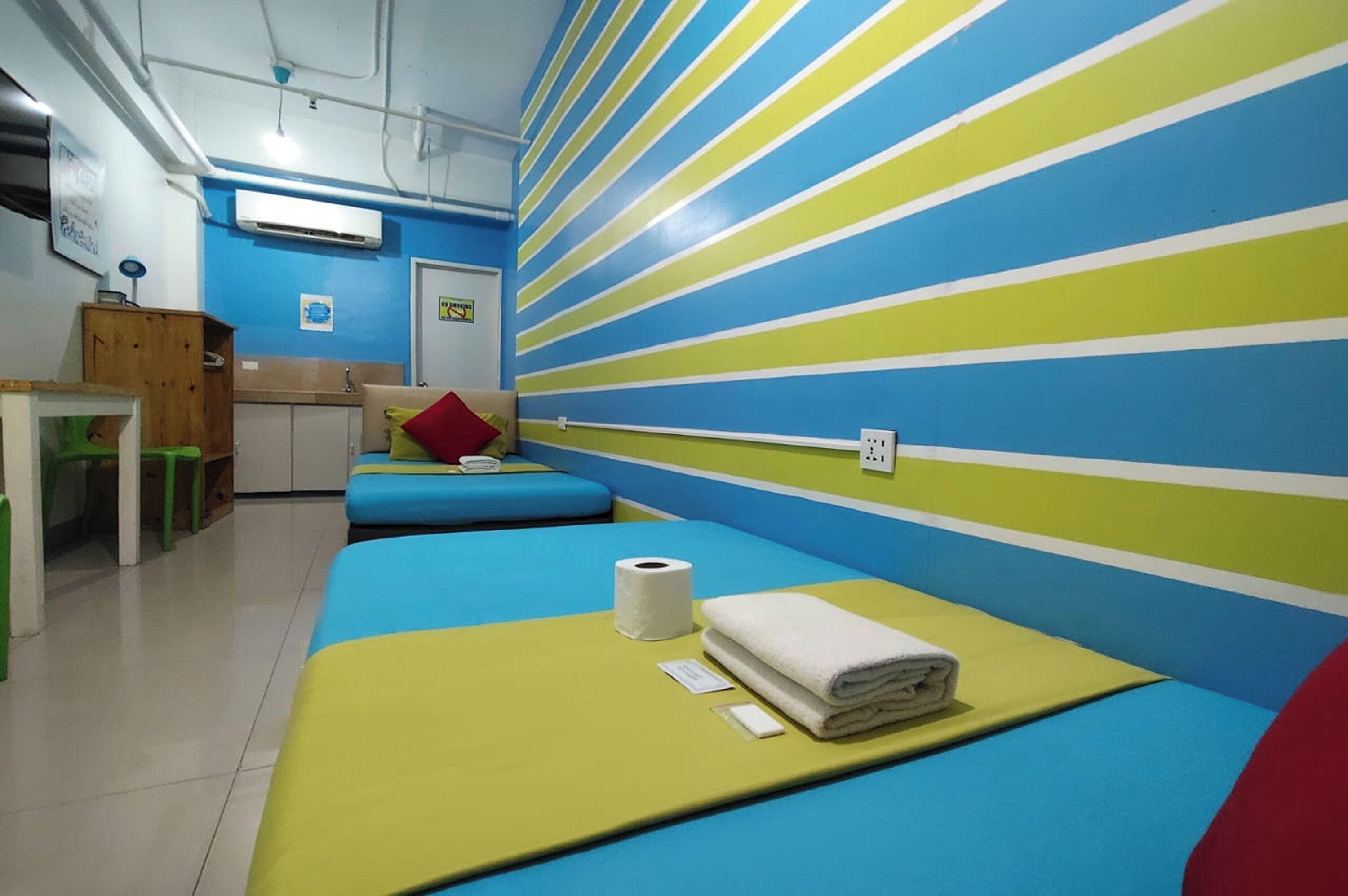 A room with striped walls, a bed with a yellow mattress, a red pillow, a white towel, and a toilet paper roll.