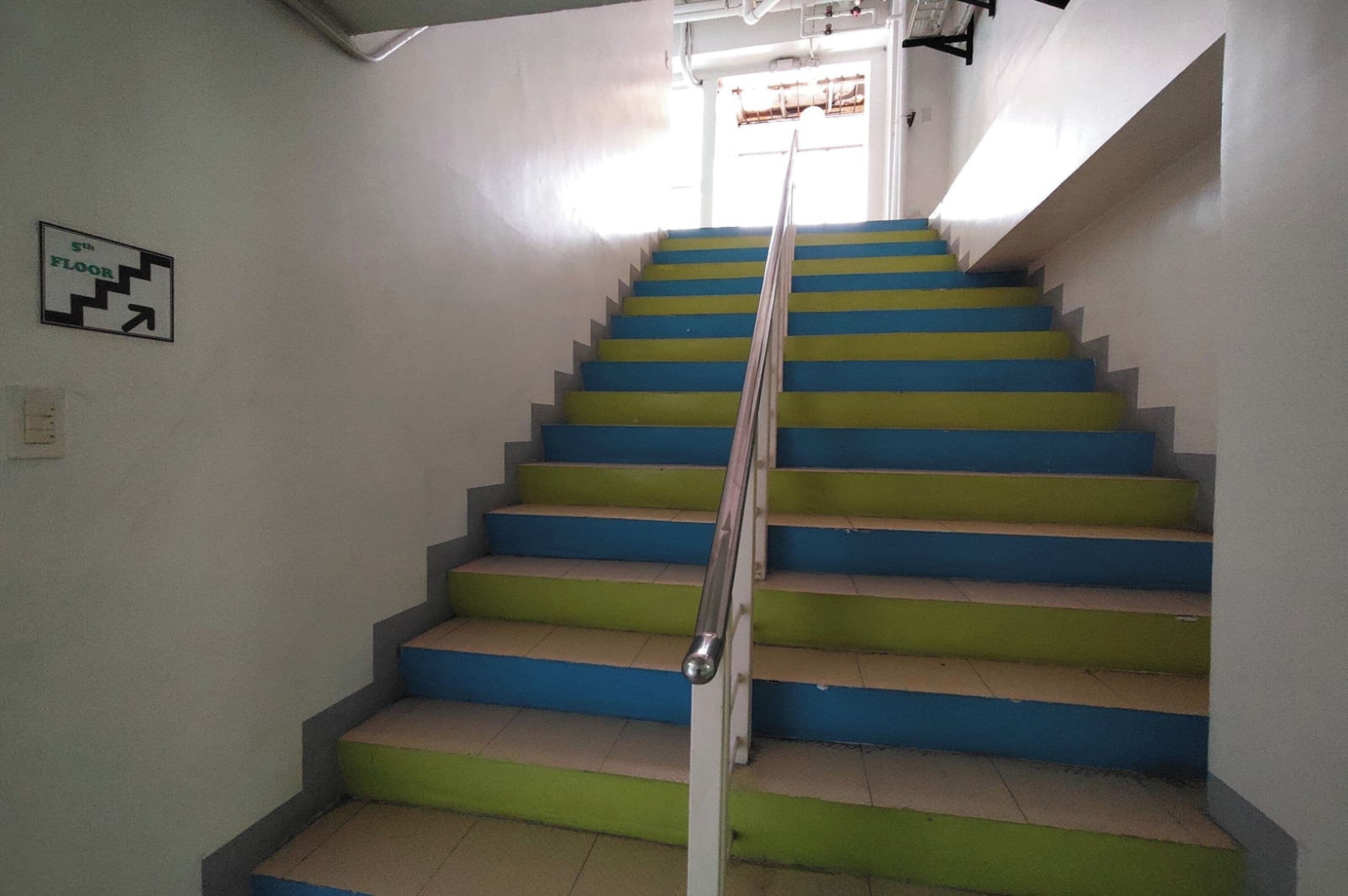 A staircase with alternating blue and green steps, a handrail, and a sign indicating the 5th floor.