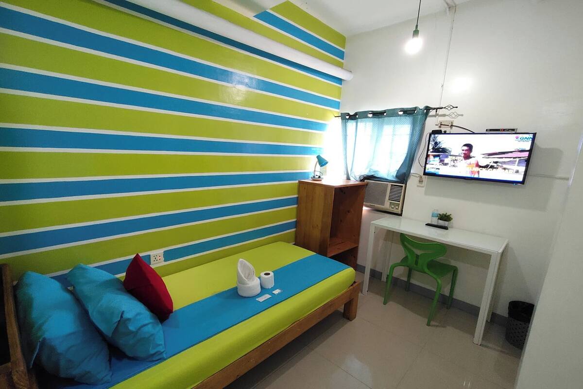 A room with a striped wall, a bed with pillows, a desk with a chair, and a television.