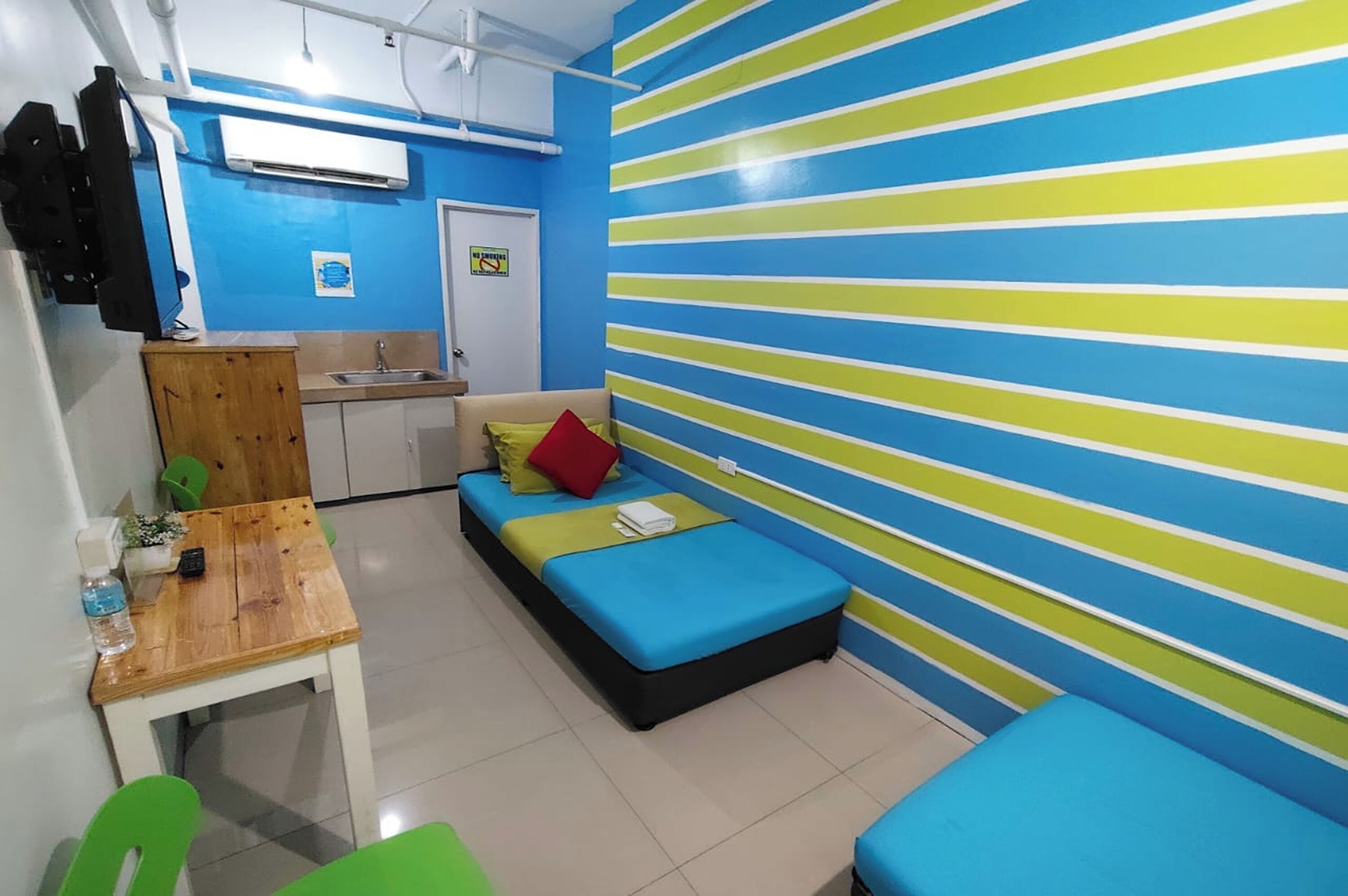 A small, brightly colored room with a bed, a table, and a kitchenette.