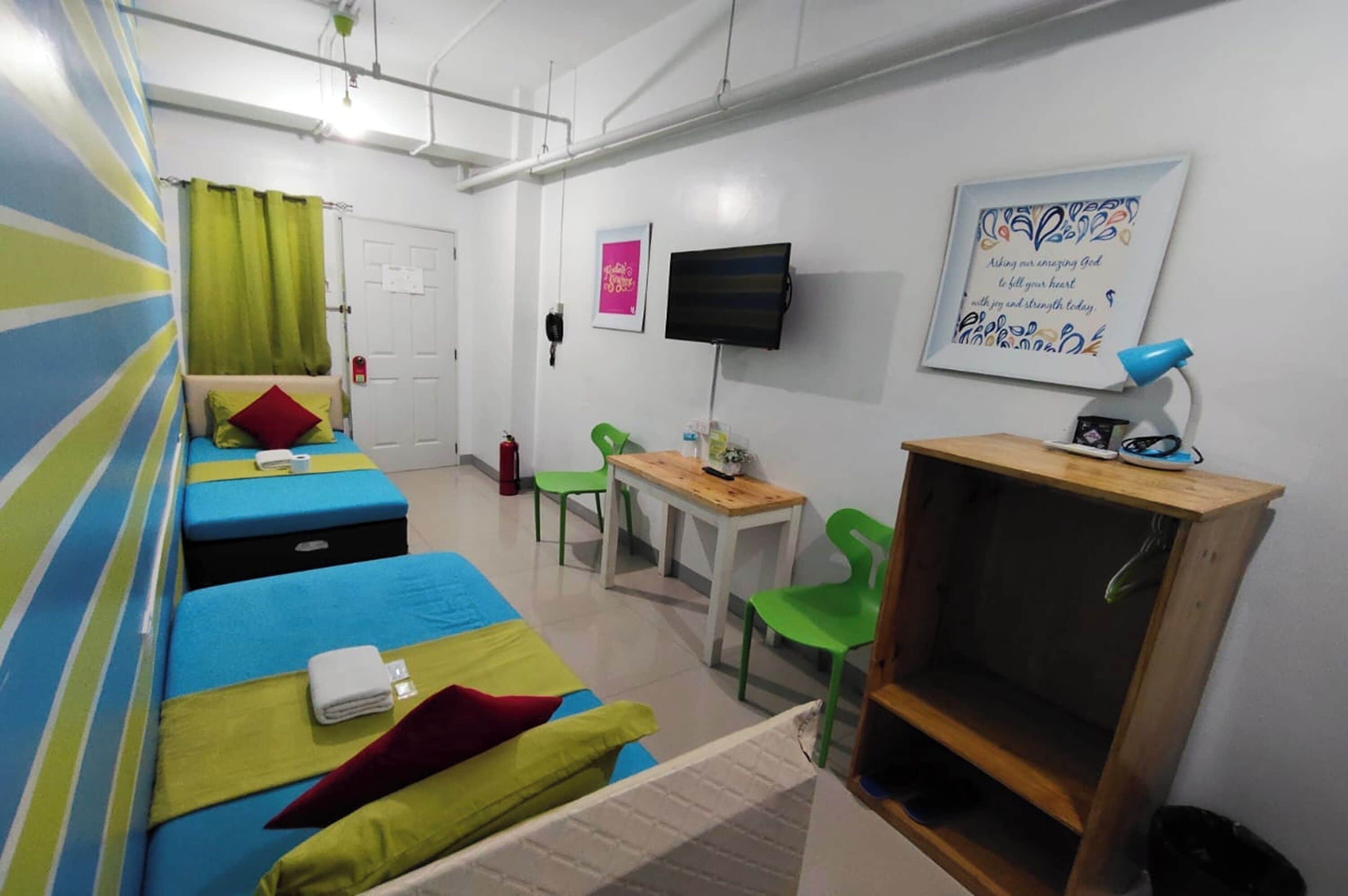 A room with two beds, a desk, a chair, a television, and a wall-mounted shelf.
