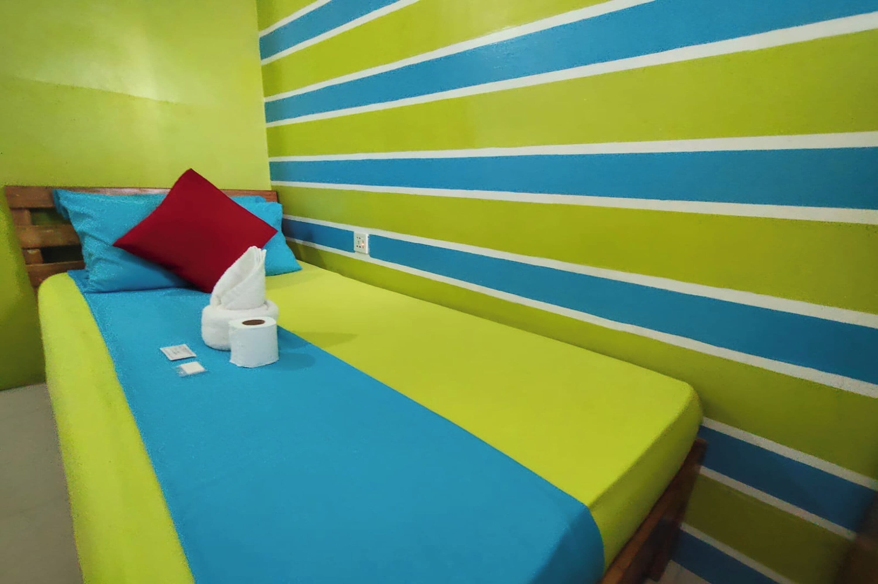 Una cama individual con una pared azul y verde a rayas, un cabecero de madera y una almohada roja.