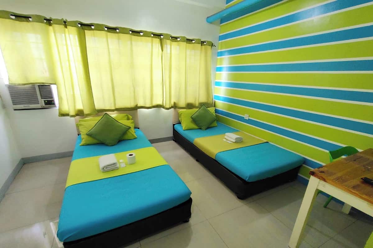 A hotel room with two beds, each with a blue mattress and green pillows, a small table, and a striped wall.