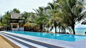 Zona de piscina con una estructura de techo de paja, palmeras y vista al mar.