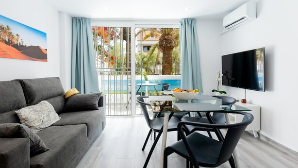 A modern living room with a grey sofa, a dining table with chairs, a flat-screen TV, and a balcony with a pool view. - 101 Playa Honda, Playa de las Americas (Playa de las Américas)