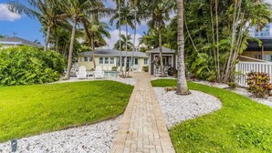 Una casa tropical con un camino pavimentado que lleva a una zona de piscina, rodeada de palmeras y exuberante vegetación.