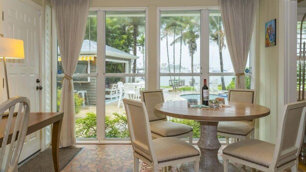Un comedor con una mesa redonda y cuatro sillas, ventanas amplias con cortinas, y vista a una piscina y mobiliario exterior.