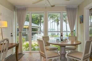 Un comedor con una mesa redonda y cuatro sillas, ventanas amplias con cortinas, y vista a una piscina y mobiliario exterior.