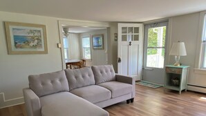 Un salon avec un canapé d’angle beige, un plancher en bois, une table à manger avec des chaises, une porte avec un panneau de verre et une petite table avec une lampe.