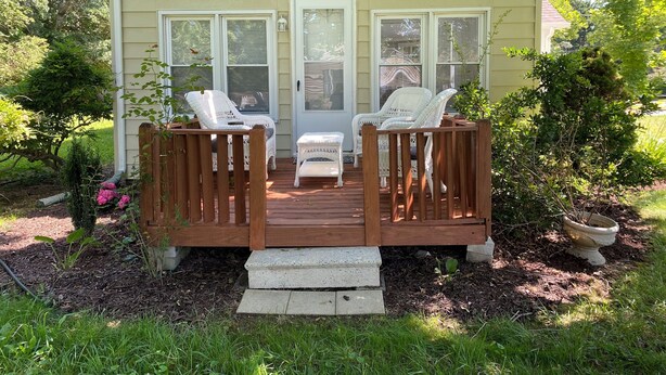 Une terrasse couverte avec deux fauteuils en osier blancs et une petite table, donnant sur une maison à l’extérieur beige et aux ornements blancs.