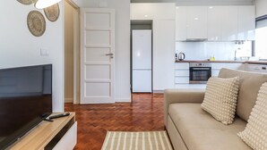 Una sala moderna con un televisor de pantalla plana, un sofá y una mesa de centro. Se observa una cocina con alacenas blancas y una estufa.