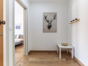 Um corredor com piso de madeira, parede branca, uma porta, uma cama ao fundo, uma mesinha branca e uma estante com plantas.
