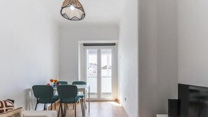 Ein modernes Wohnzimmer mit einer Couch, einem Esstisch mit Stühlen, einem Fernseher auf einem Schrank und einem großen Fenster mit Blick auf Gebäude.