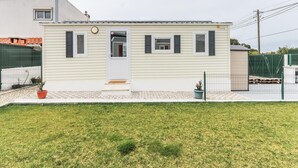 Ein eingeschossiges, helles Gebäude mit einer weißen Tür und schwarzen Fensterläden, umgeben von einem grünen Zaun und einer kleinen Rasenfläche.