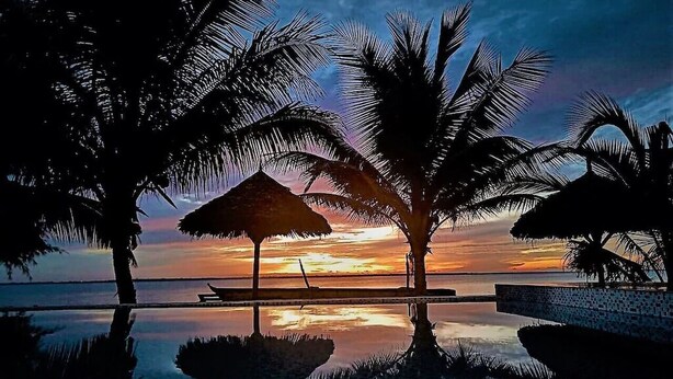 Ein friedlicher Sonnenuntergang mit Palmen und einem Reetdachschirm am Ufer eines spiegelnden Pools.